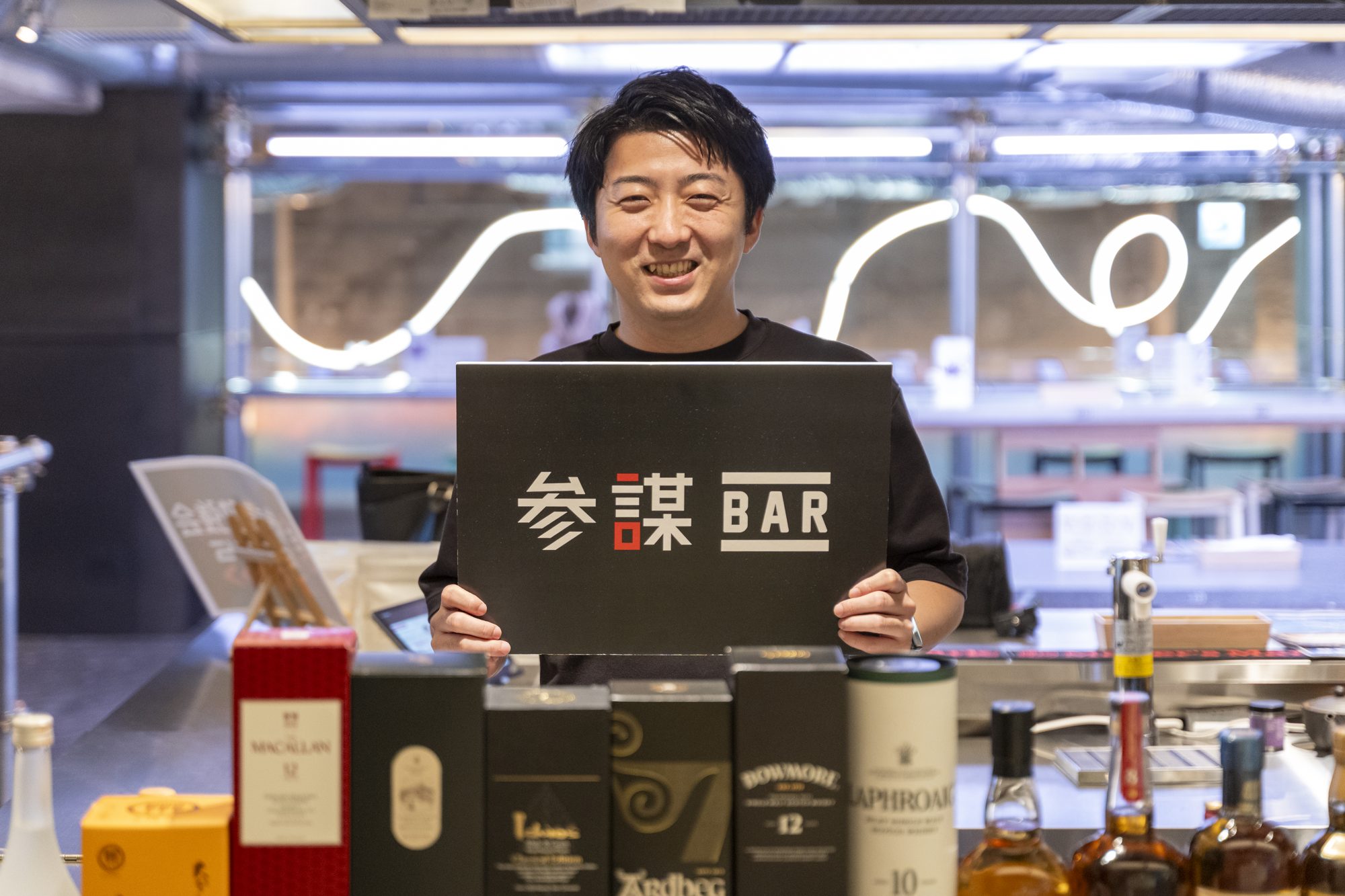 参謀BAR 新宿のカウンターで参謀BARのボードを手に笑顔を見せる株式会社RASHISA代表・岡本翔さん